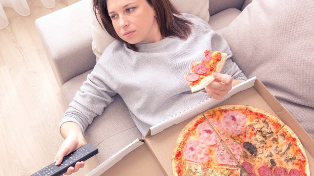 woman eating pizza and holding phone laying on sofa at home